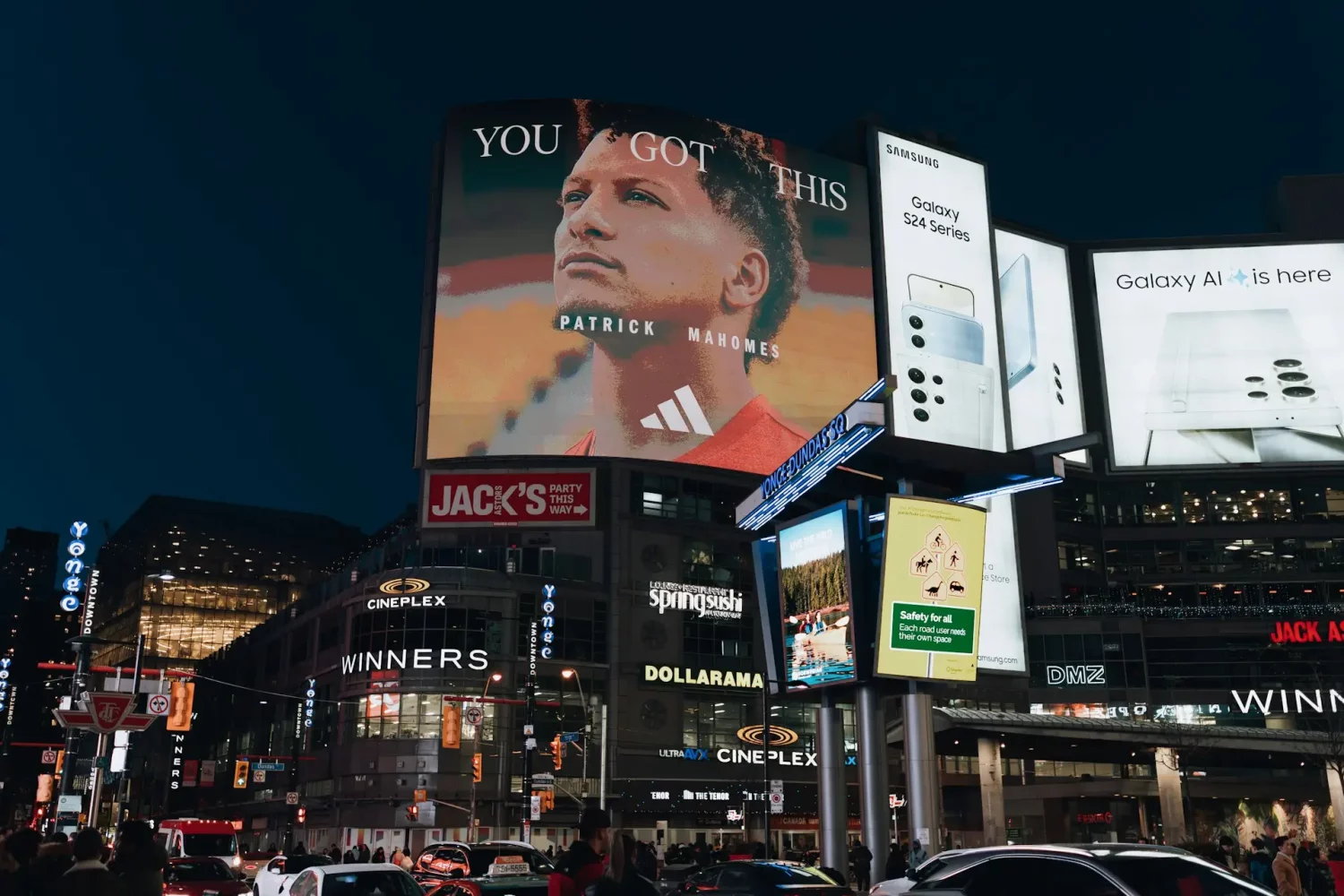 Nocny plac miejski z dużym billboardem Patricka Mahomesa i reklamami Samsunga. Przykład, jak reklamować firmę w centrum miasta.